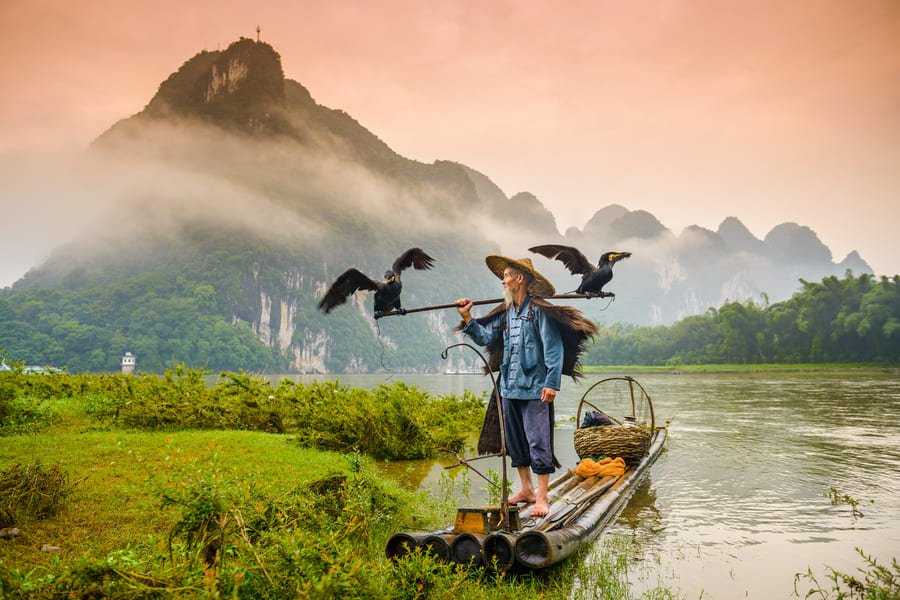 Pescador de cormorán sobre una lancha en el rio Yulong en Yangshuo con montañas de fondo